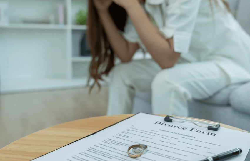 A distressed woman sitting on a sofa and divorce documents and wedding rings placed on a table.